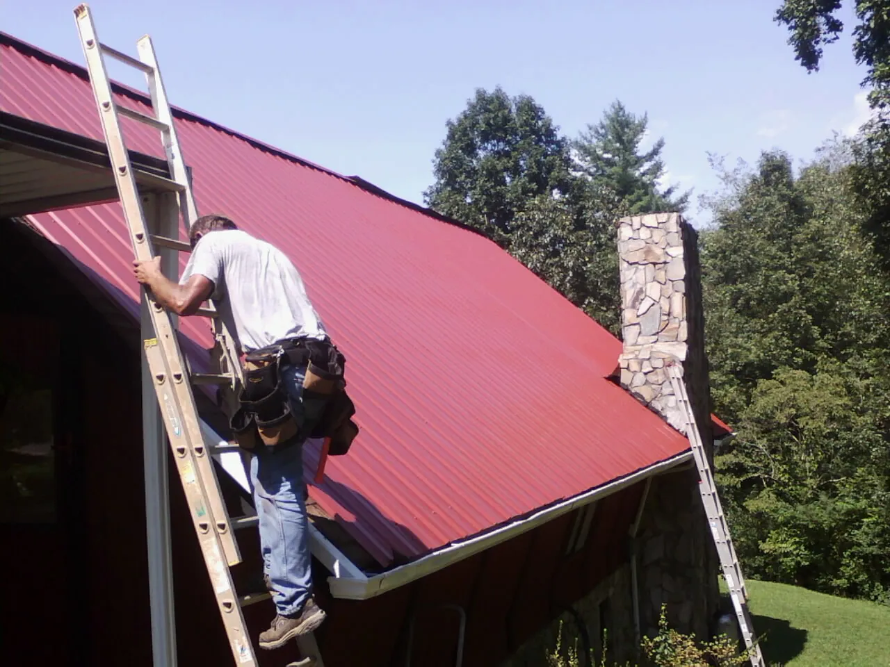 Roofer on a ladder beside a red metal roof and stone chimney — Soffit & Fascia Repair by Roofing Brothers in Cassville